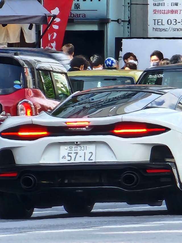 A white sports car with distinctive rear lights is parked on a busy street, surrounded by other vehicles and pedestrians