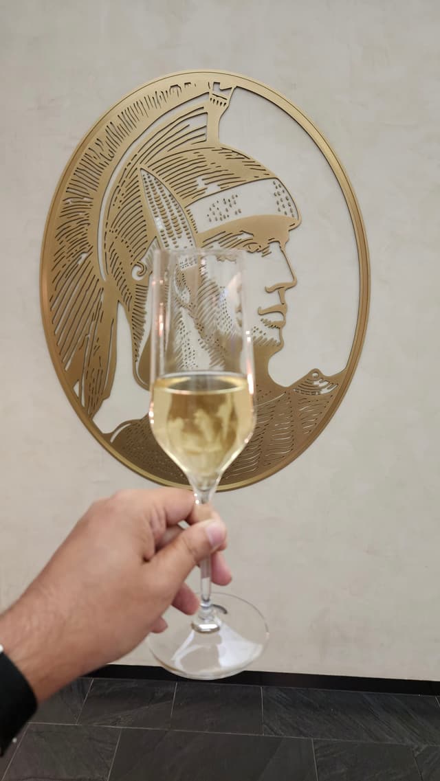 A hand holding a glass of champagne in front of a gold-colored wall decoration featuring a profile of a person wearing a hat