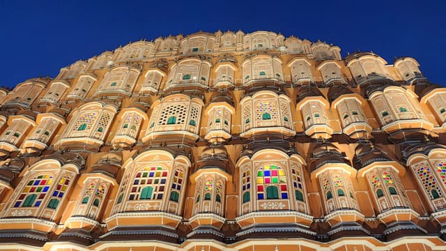 An ornate, multi-story building with numerous small windows, each adorned with intricate latticework and colorful glass, illuminated against a deep blue evening sky