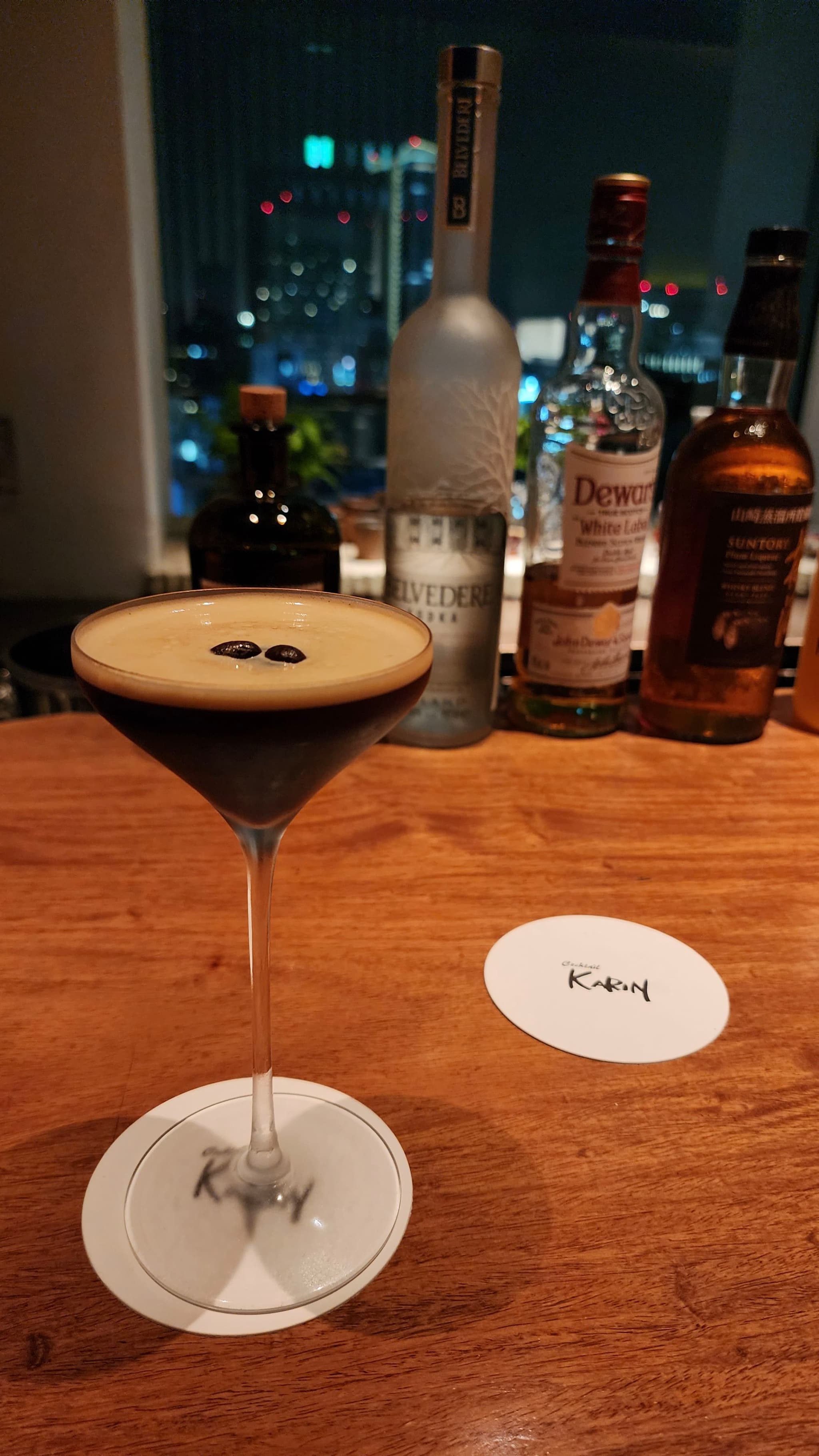 A cocktail in a martini glass with two coffee beans on top, placed on a wooden bar counter with various liquor bottles in the background and a cityscape visible through the window