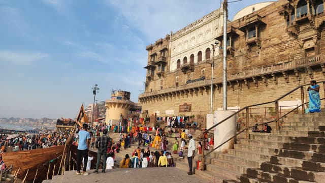 A bustling scene at the ghats of Varanasi, India, with people gathered near the steps leading to the Ganges River The architecture features historic buildings with intricate designs, and the area is lively with activity, including pilgrims and tourists The sky is clear, and the atmosphere is vibrant