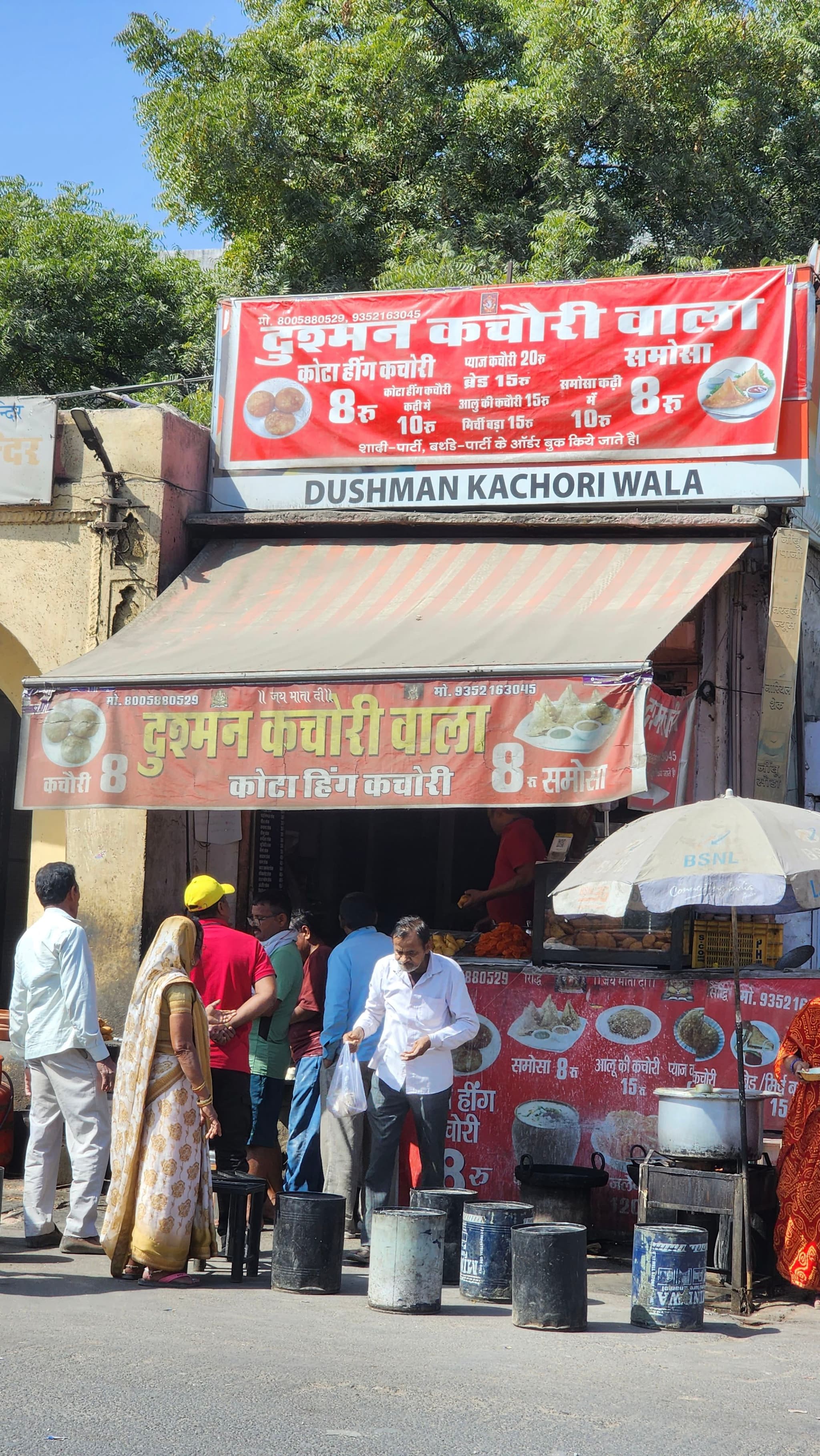 A street food stall with red signage and several people standing around, some of whom appear to be customers The stall has a canopy and various cooking utensils and containers are visible Trees and a clear blue sky are in the background