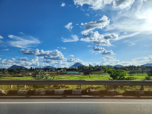 A scenic landscape featuring a clear blue sky with scattered clouds, green fields, distant mountains, and a road with a guardrail in the foreground