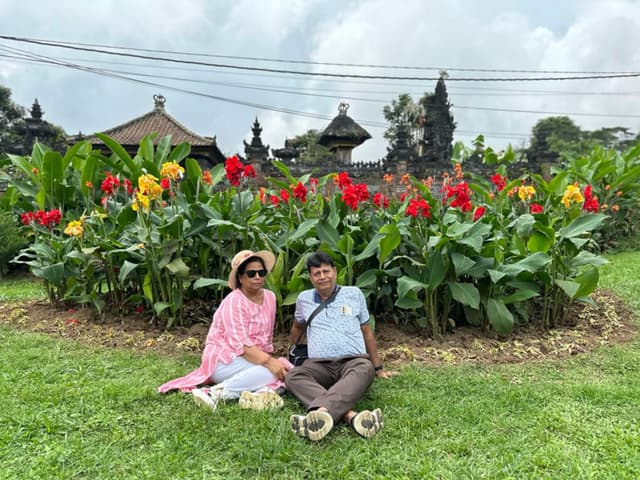 A couple sits on the grass in front of a vibrant flower bed with red and yellow blooms, with traditional Balinese temple structures visible in the background