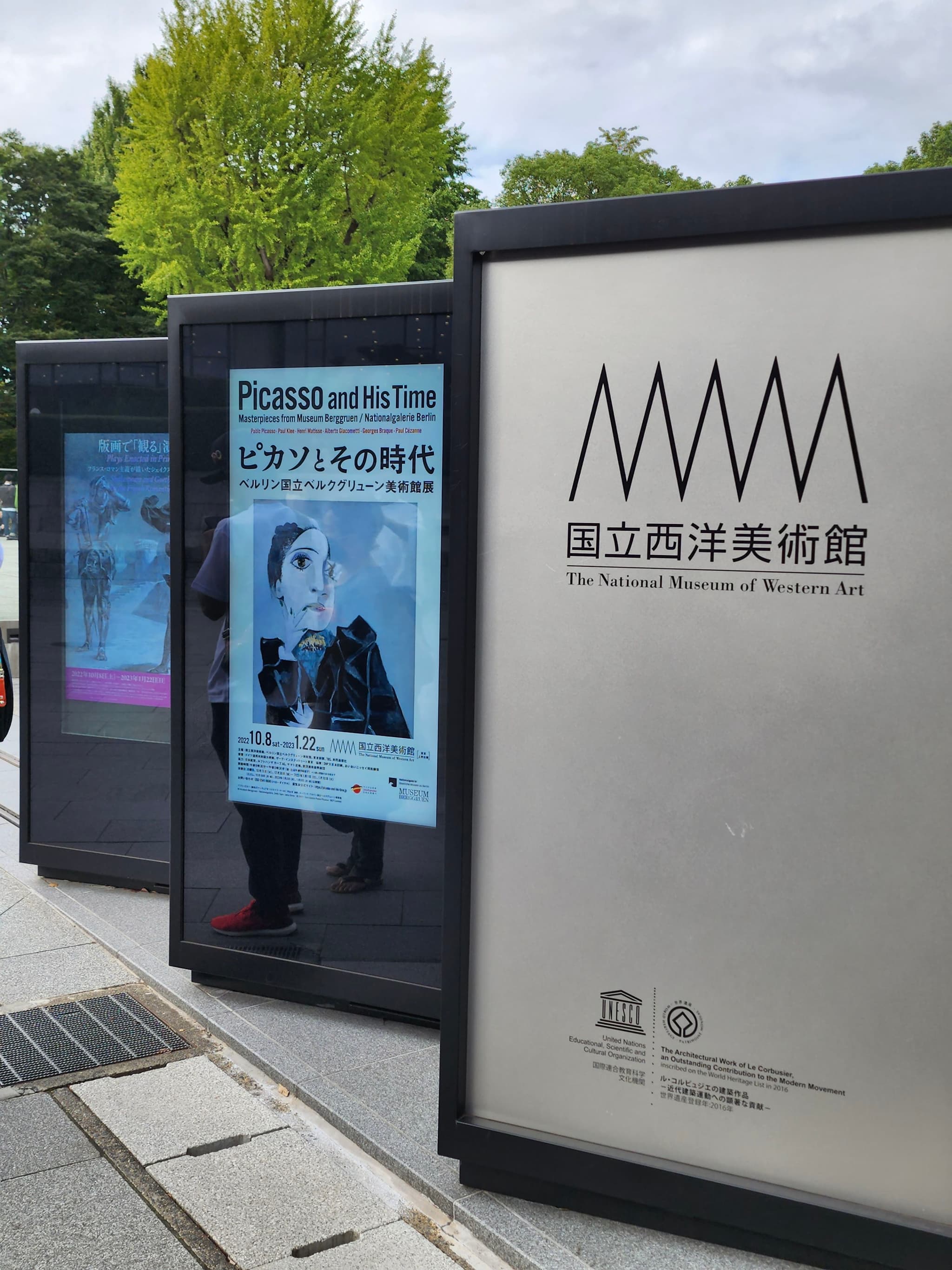 A series of outdoor posters, including one for the National Museum of Modern Art in Tokyo, displayed on black stands along a sidewalk with trees in the background