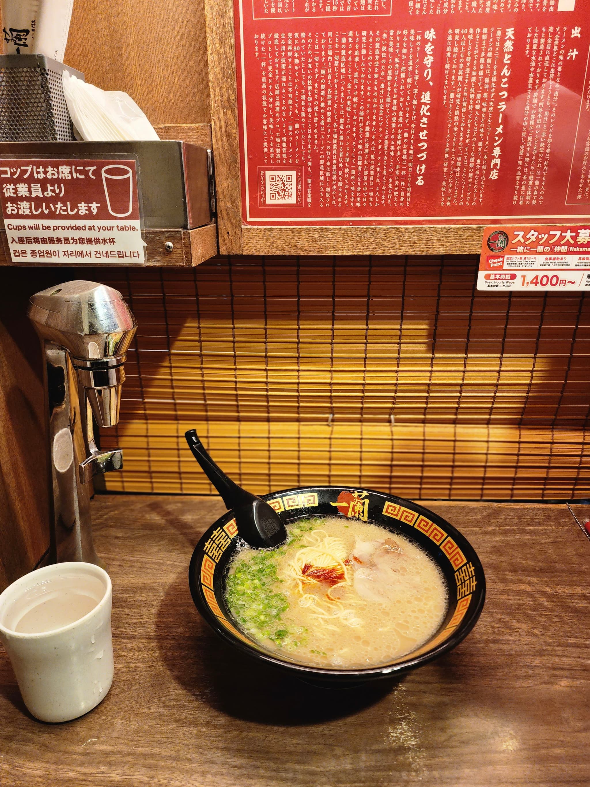 A bowl of ramen with a spoon, a cup, and a tissue dispenser on a wooden table in a small dining booth