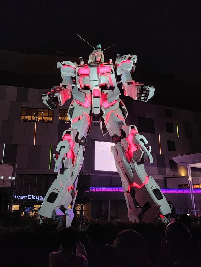 A large, illuminated robot statue with pink and white lights stands prominently in front of a building at night, surrounded by onlookers