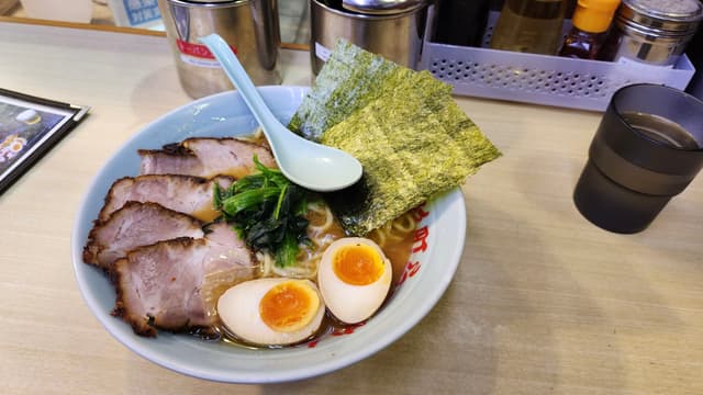 A bowl of ramen with sliced pork, boiled eggs, seaweed, and vegetables, accompanied by a spoon, on a table with condiments and a glass of water in the background