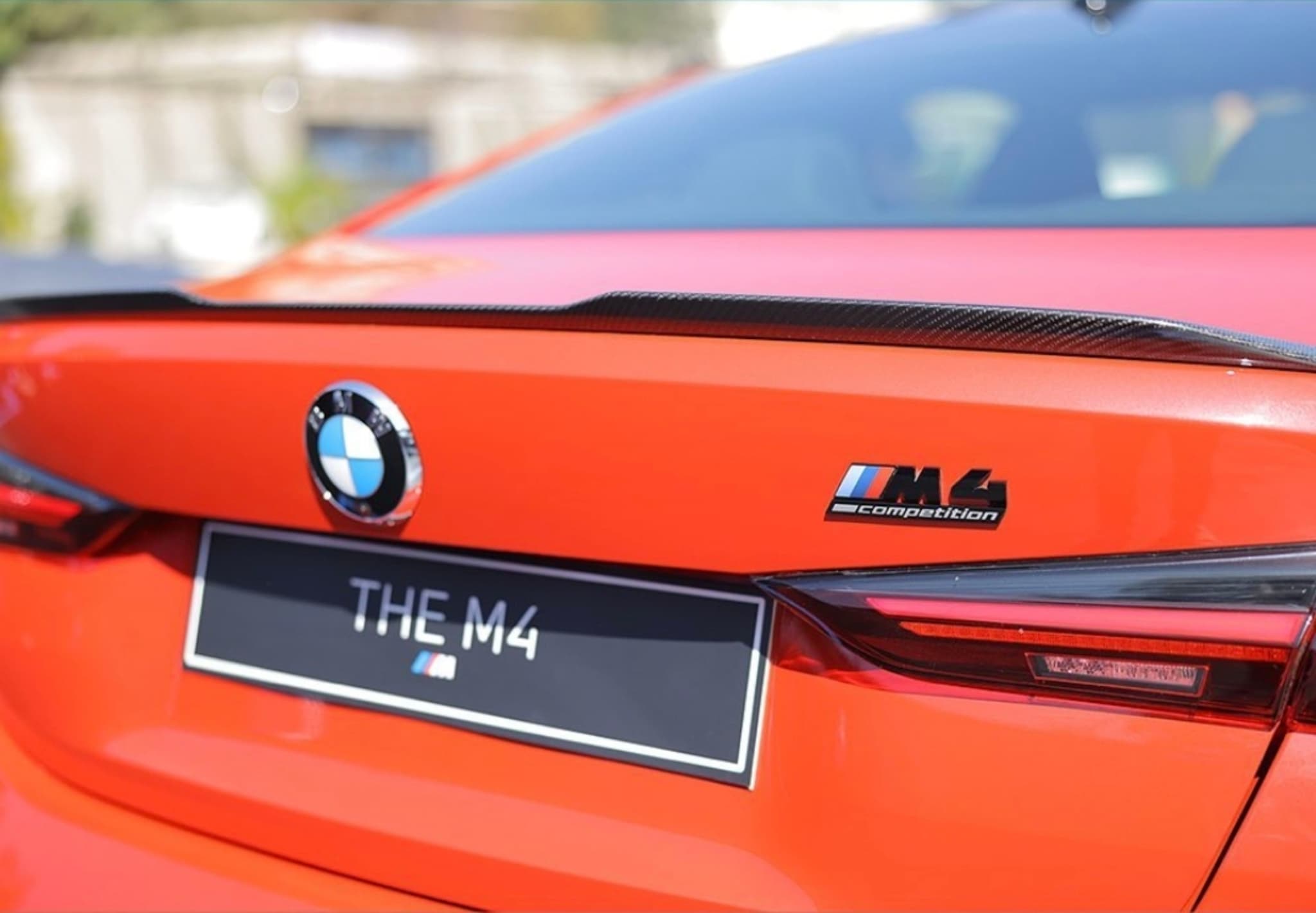 The rear end of an orange BMW M4, featuring the BMW logo, M4 badge, and a license plate reading THE M4