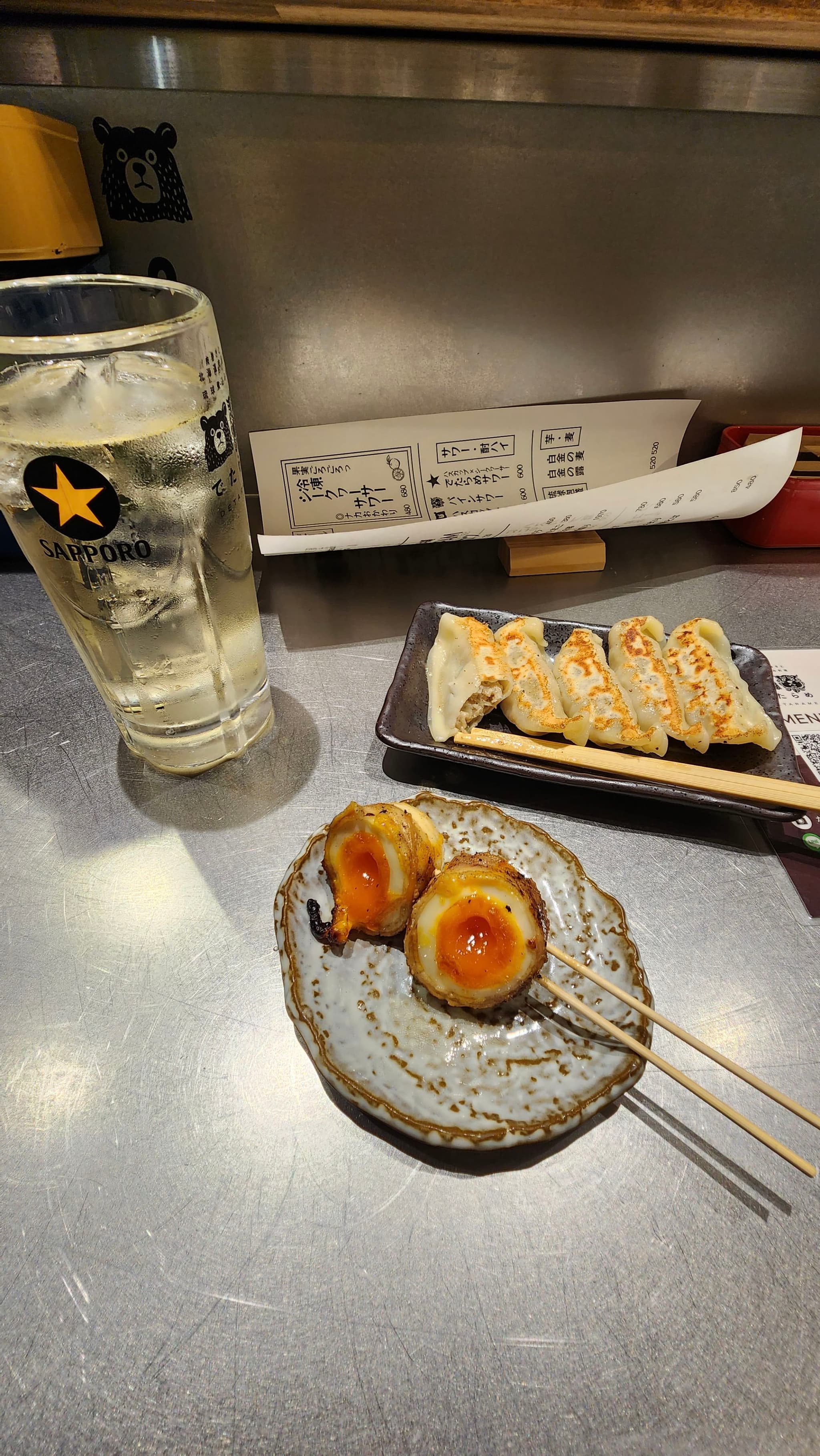 A glass of beer, a plate of gyoza dumplings, and skewered food items on a small plate are arranged on a metallic table