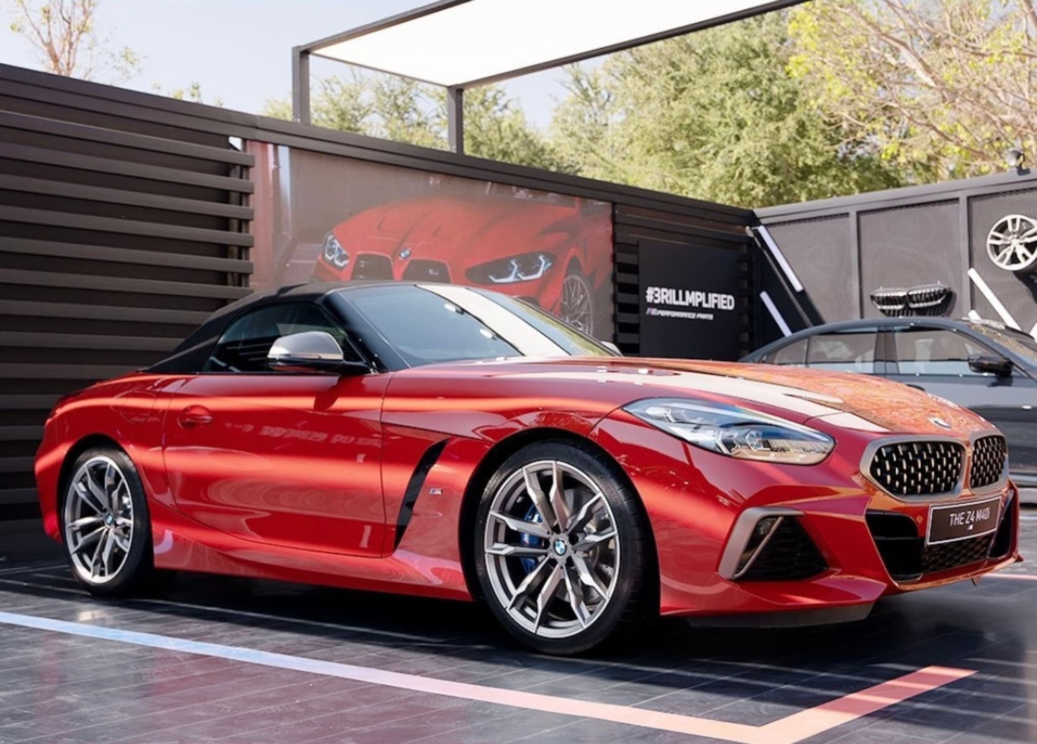 A sleek, red sports car with a black roof is parked in a modern, outdoor setting with other vehicles and automotive-themed decor in the background