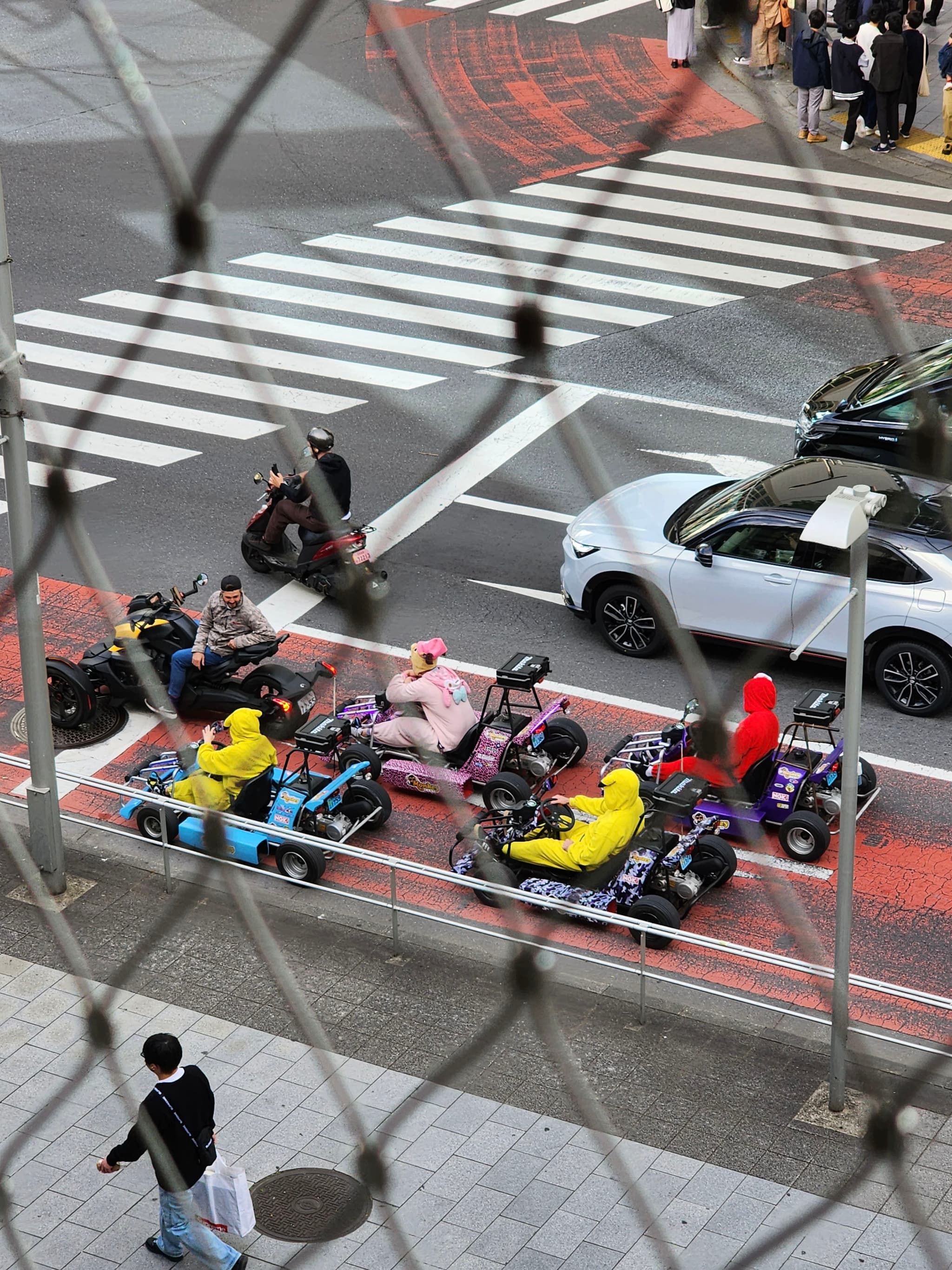 A group of people in colorful costumes riding go-karts on a city street, viewed from above through a chain-link fence