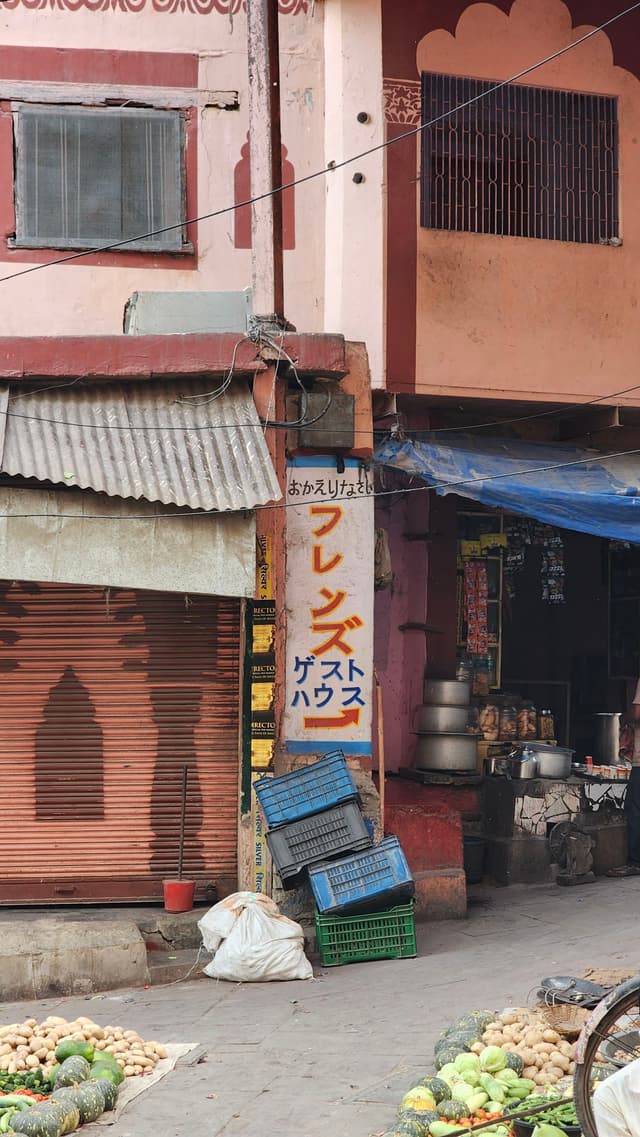 A narrow street with a small shop displaying a sign in a japanese kana script, surrounded by crates and produce The shop has a corrugated metal awning and a partially closed rolling shutter Nearby, there are baskets of vegetables, indicating a market area