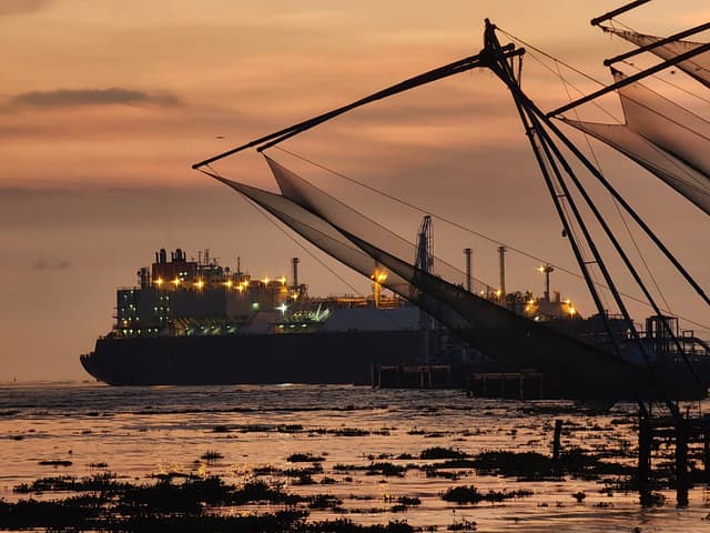 A large cargo ship illuminated with lights is seen in the background, while traditional fishing nets are silhouetted against a sunset sky in the foreground