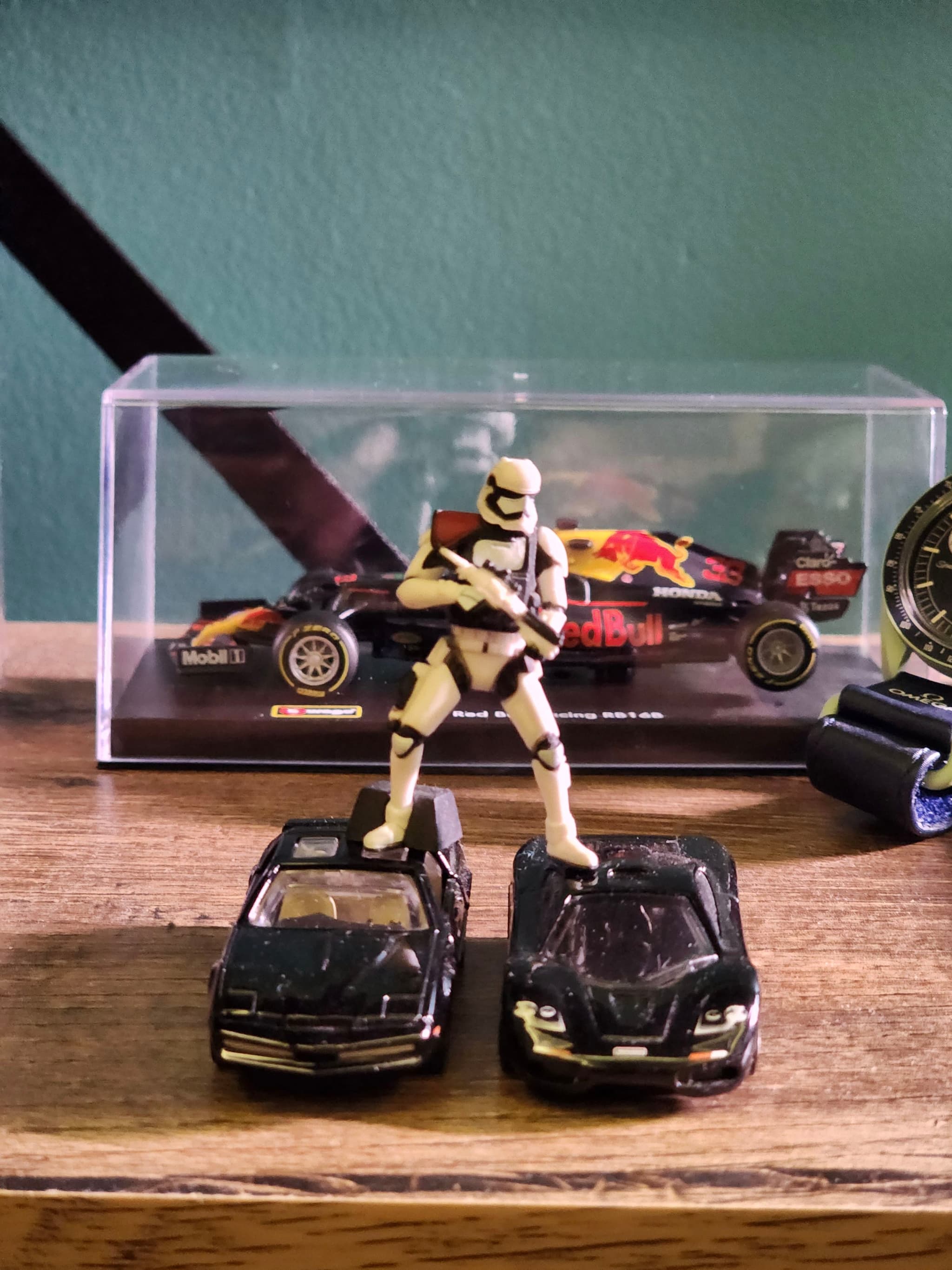 A toy figure resembling a stormtrooper is standing on two black toy cars, with a model car displayed in a clear case in the background