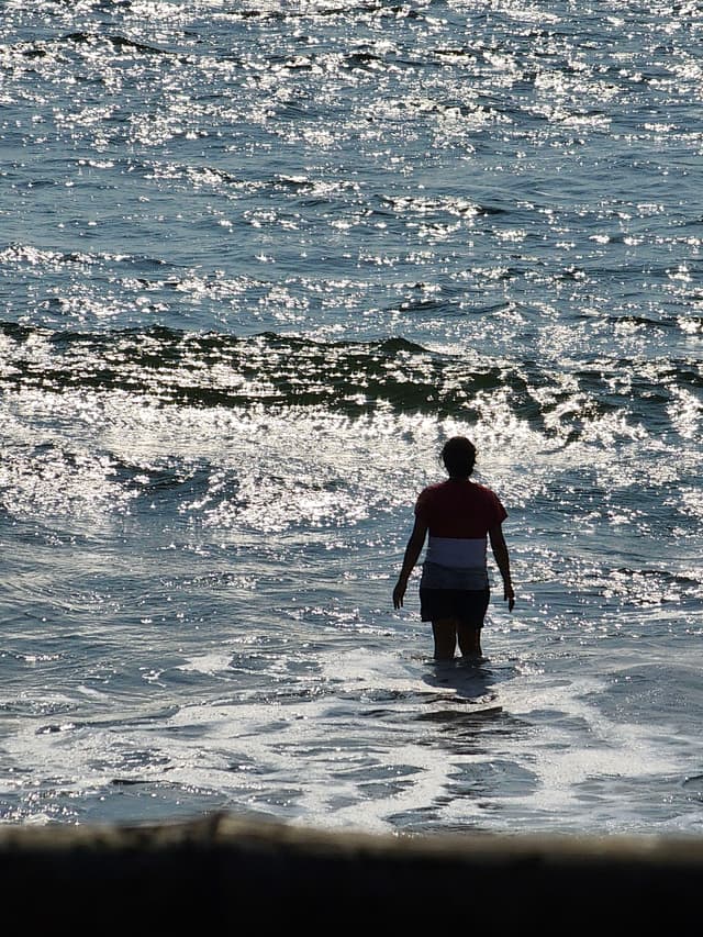 A person standing in the shallow water of a beach, facing the ocean with waves crashing around them