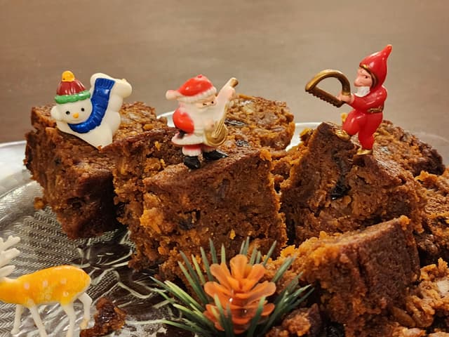 Several pieces of fruitcake are decorated with small figurines, including a snowman, Santa Claus, and an elf, along with a small plastic deer and a pinecone