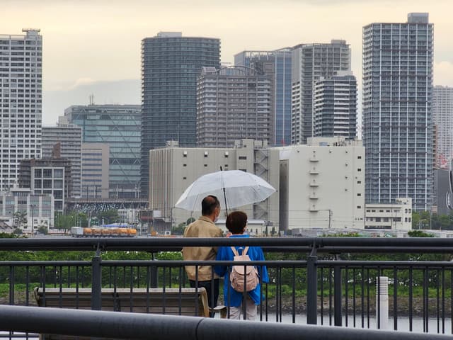 Two people standing on a bridge, one holding an umbrella, with a cityscape of tall buildings in the background