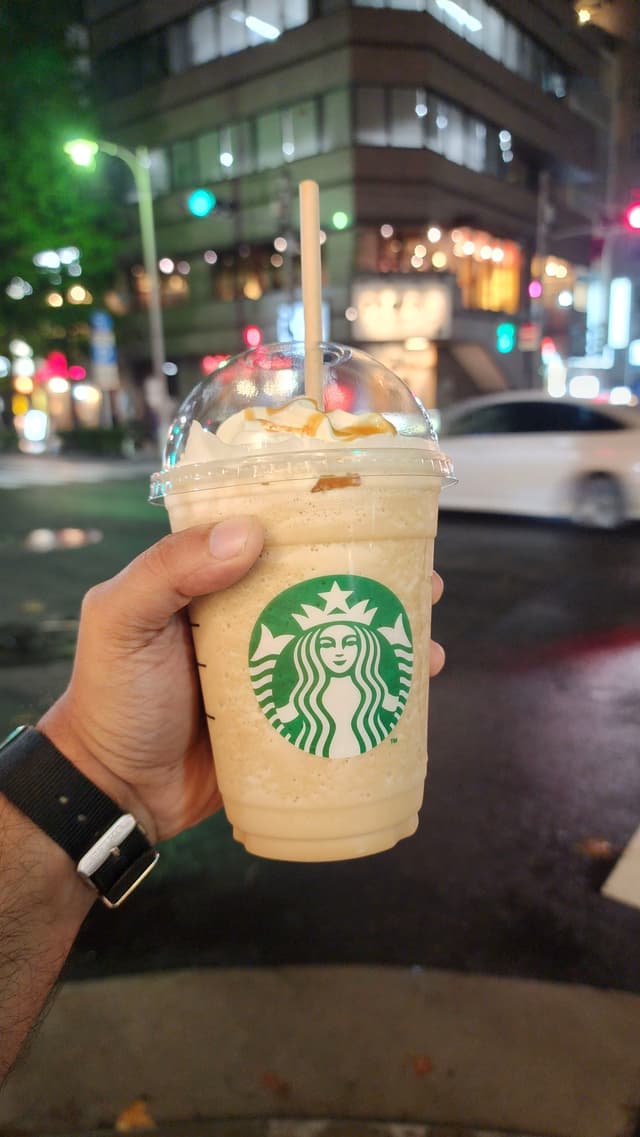 A hand holding a Starbucks frappuccino with whipped cream and caramel drizzle, against an urban nighttime background with blurred lights and buildings