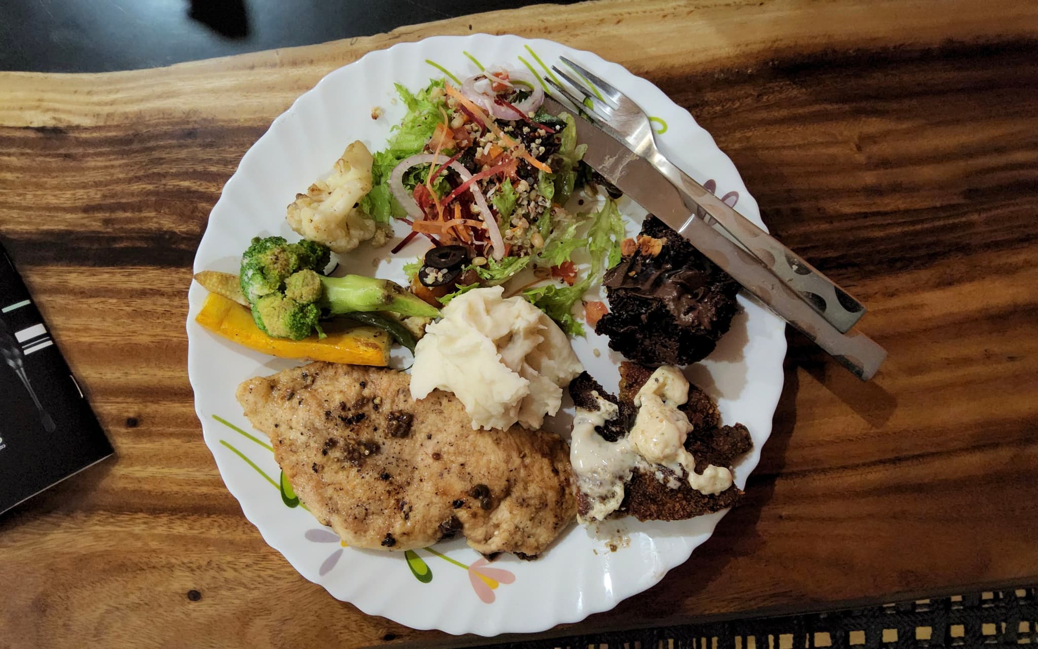 A plate of food featuring grilled chicken, a salad with mixed greens and red onions, roasted vegetables including broccoli and carrots, and a dollop of hummus A fork and knife rest on the plate, which is placed on a wooden table