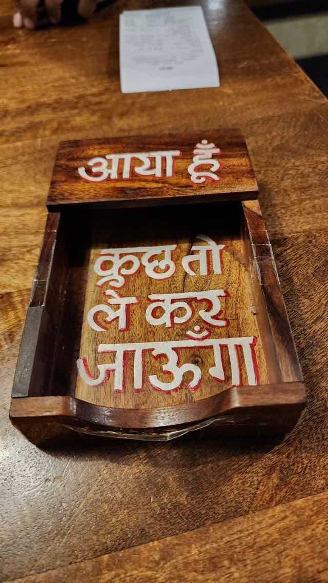 A wooden box with intricate carvings and white painted text in a script, placed on a wooden surface