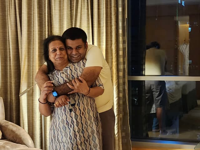 A man hugging a woman from behind in a warmly lit room with curtains and a window reflecting their image