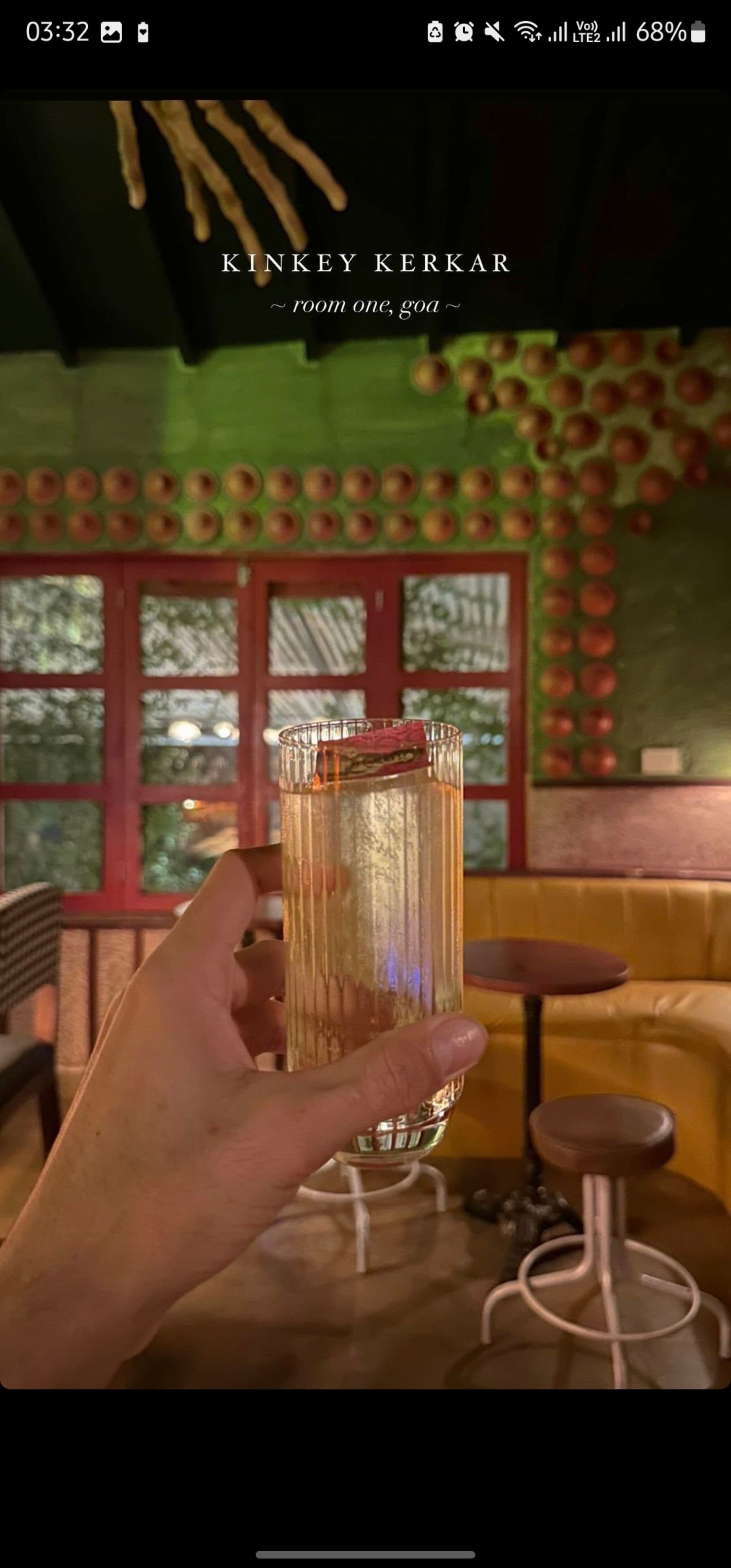 A hand holding a glass of a light-colored beverage garnished with a slice of fruit, set against the backdrop of a cozy, dimly lit café with green walls, red window frames, and a seating area with round tables and chairs