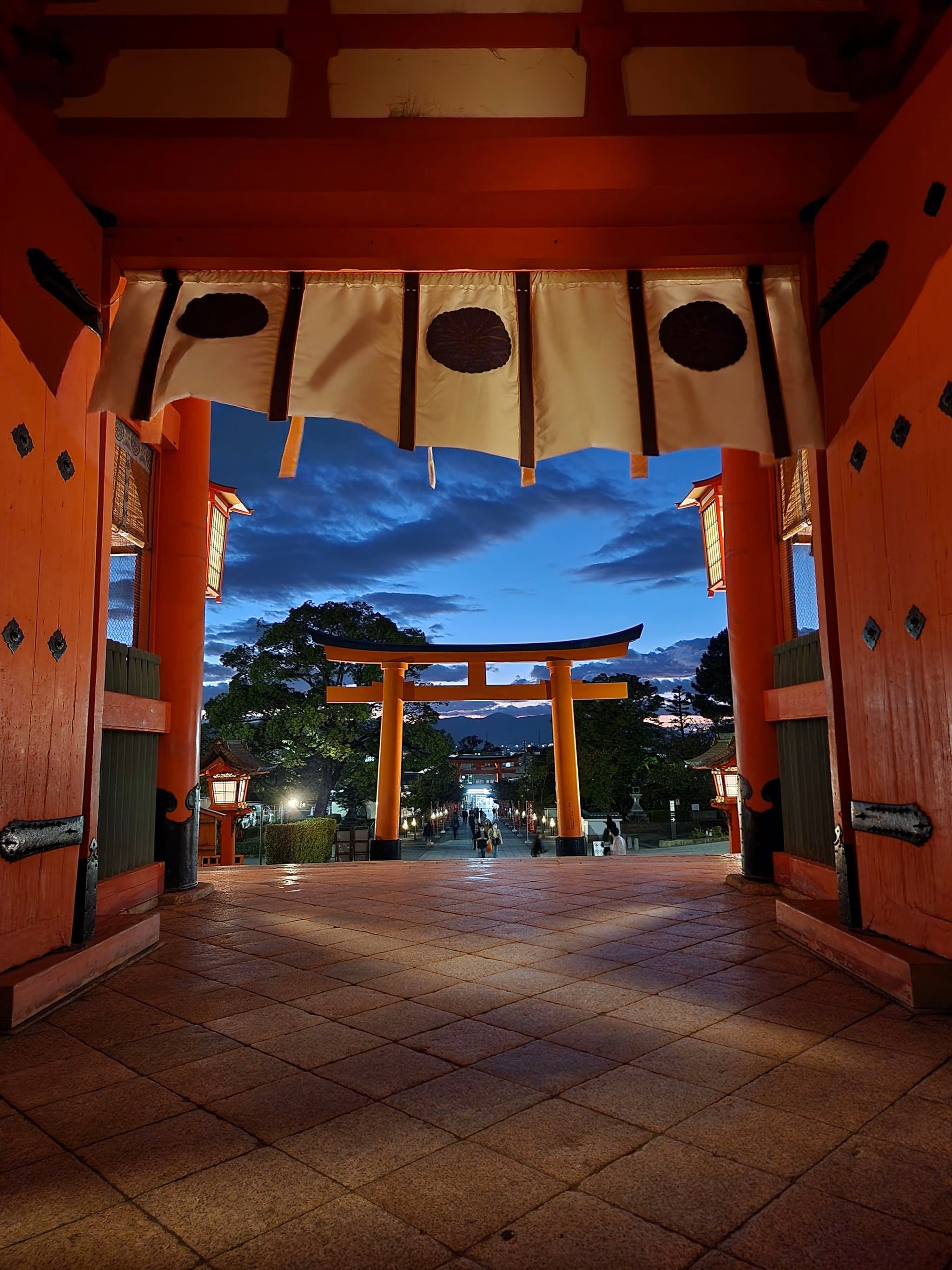 A traditional Japanese torii gate framed by the entrance of a shrine, with a view of the evening sky and surrounding trees