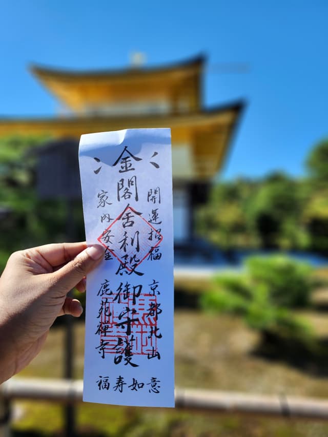 A hand holding a piece of paper with Japanese calligraphy in front of a traditional Japanese building with a golden roof, set against a clear blue sky and lush greenery