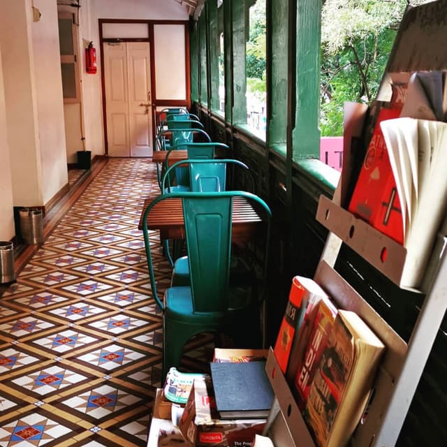 A cozy indoor café with a row of green metal chairs and wooden tables along a windowed wall The floor features a colorful geometric tile pattern A bookshelf on the right holds various books and magazines Natural light filters through the windows, creating a warm and inviting atmosphere