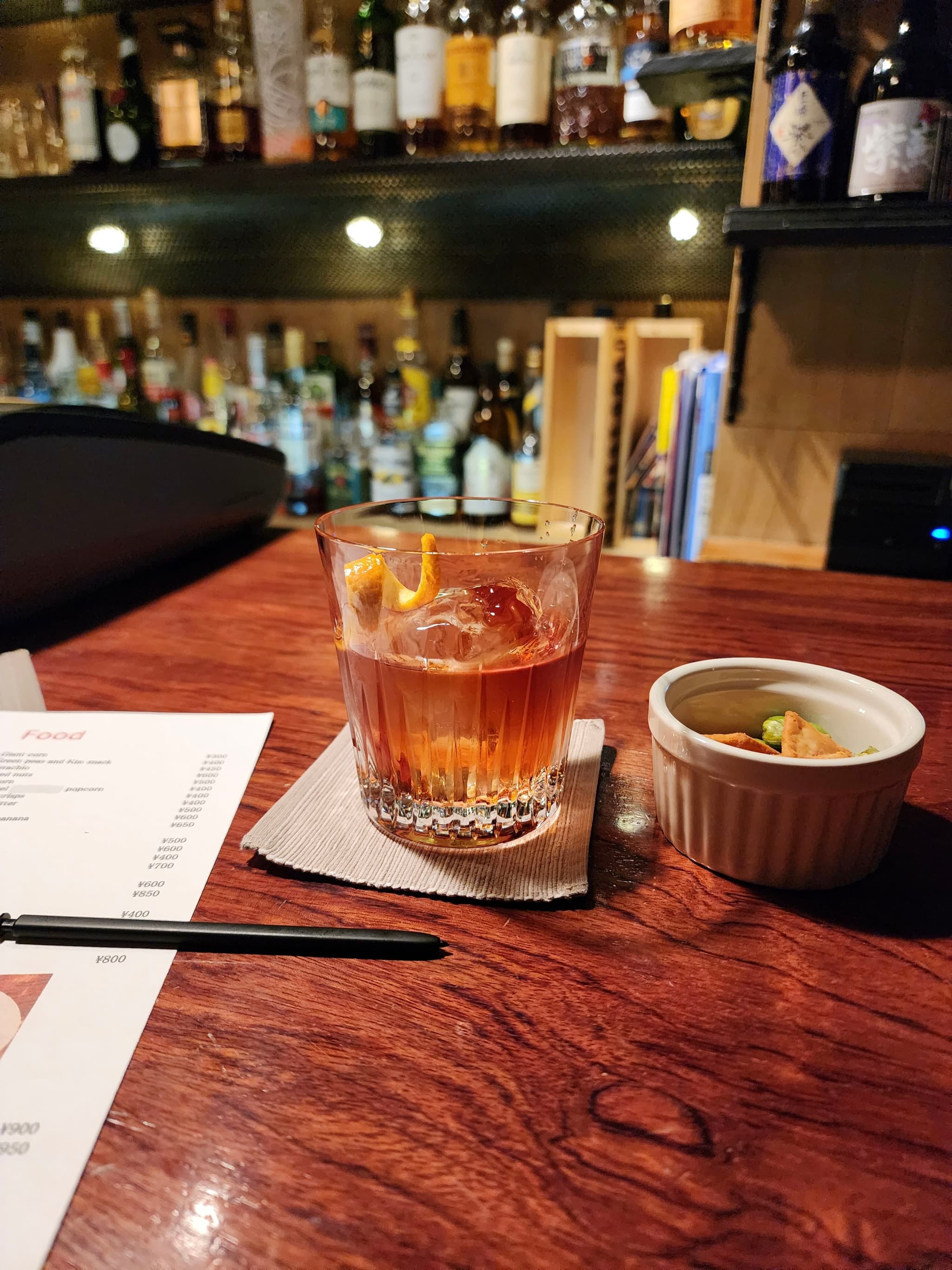 A cocktail and a small dish on a wooden bar counter, with a menu and a pen nearby, and a well-stocked bar in the background