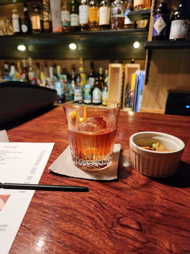 A cocktail and a small dish on a wooden bar counter, with a menu and a pen nearby, and a well-stocked bar in the background