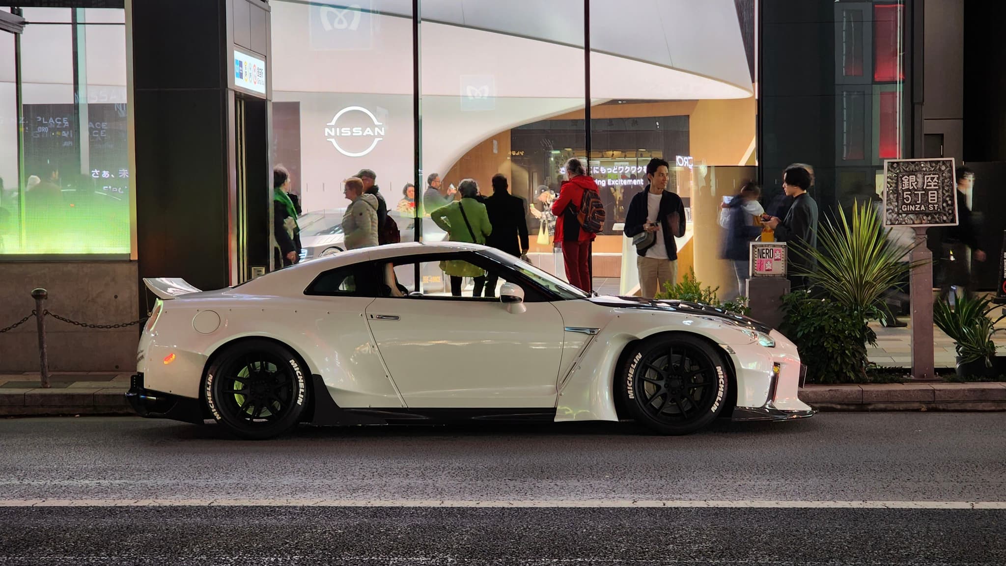 A white sports car is parked on the street in front of a building with large windows, through which several people can be seen inside