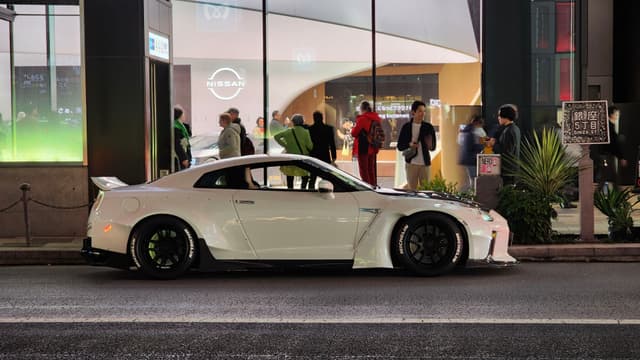 A white sports car is parked on the street in front of a building with large windows, through which several people can be seen inside