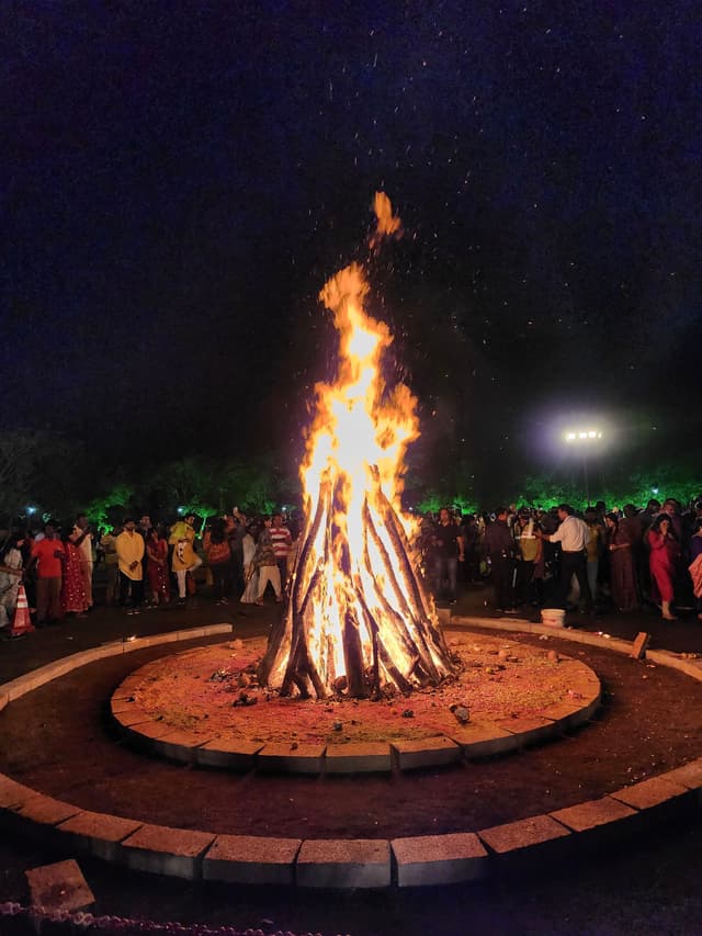 A large bonfire surrounded by a crowd of people at night, with a circular stone boundary around the fire