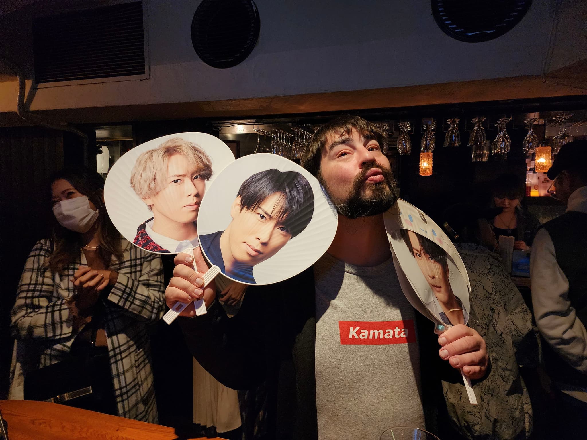 A person holding three fan signs with different faces, standing in a dimly lit room with other people in the background