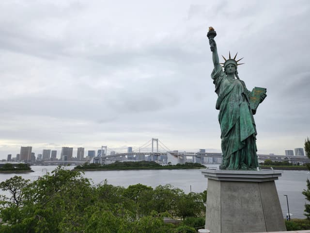 A replica of the Statue of Liberty stands prominently in the foreground with a cityscape and a bridge visible in the background, under a cloudy sky