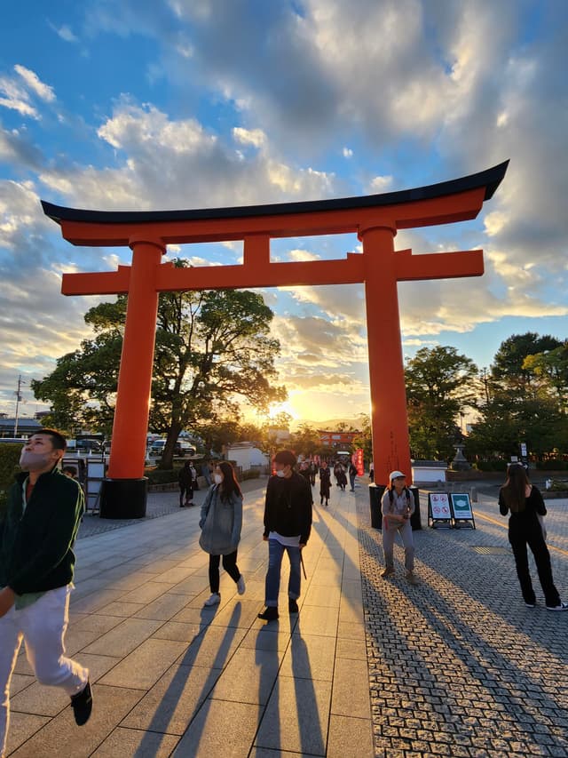 A large red torii gate stands prominently against a backdrop of a setting sun, with people walking beneath it and long shadows cast on the ground The sky is partly cloudy, and trees are visible in the background