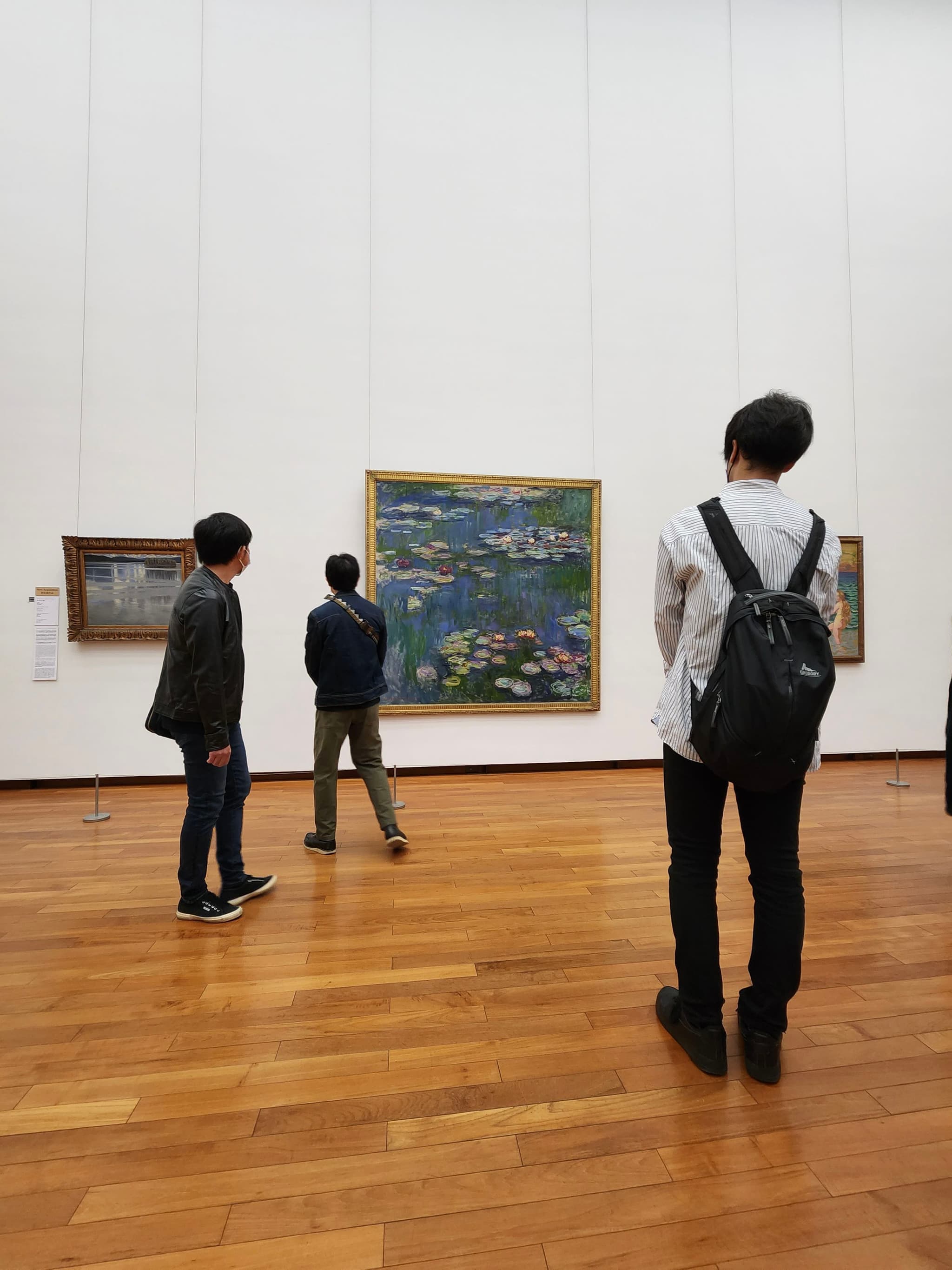 Three people observing paintings in an art gallery with wooden floors and white walls