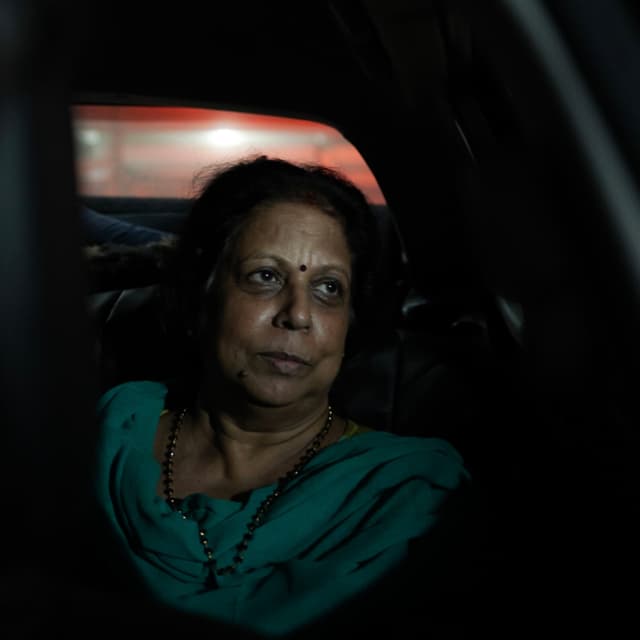 A woman in a green outfit is sitting in a dimly lit car, looking out the window with a contemplative expression