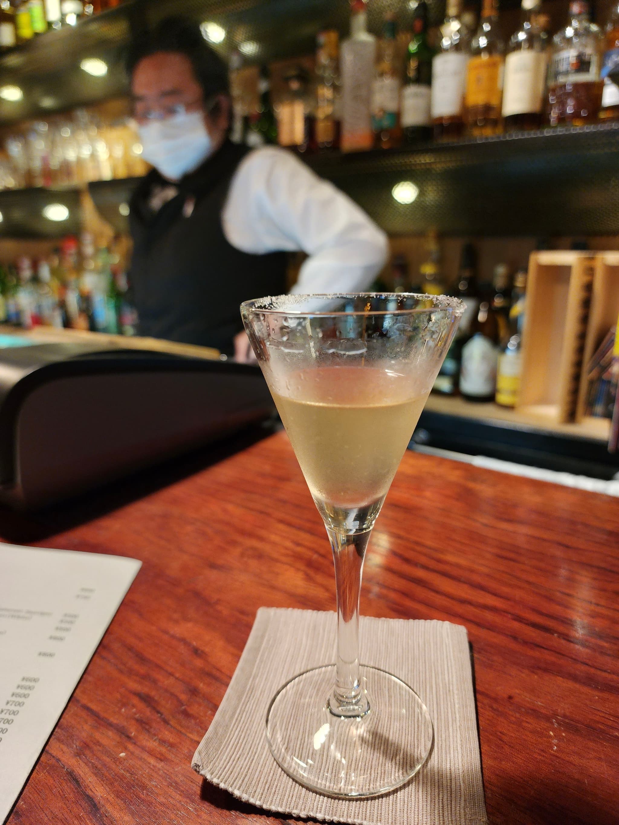 A cocktail in a martini glass on a wooden bar counter with a napkin underneath, a bartender in the background, and a menu partially visible on the right