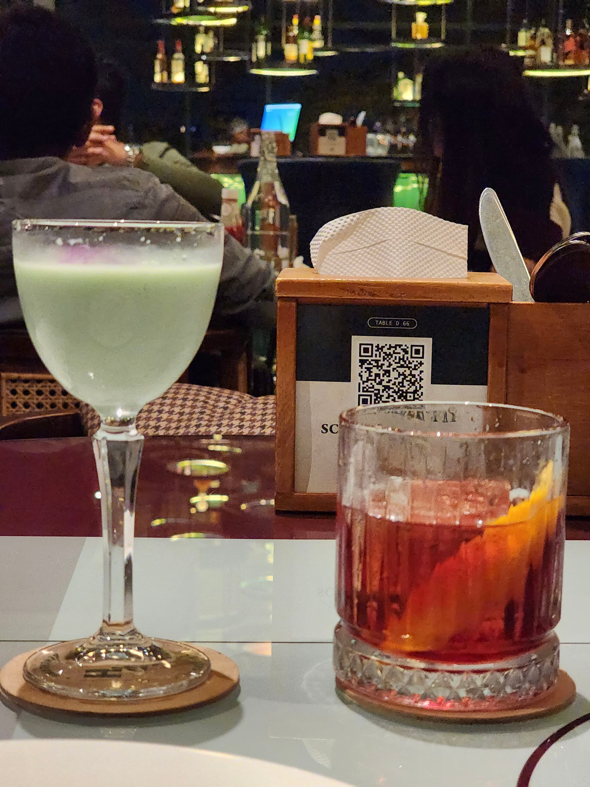 Two cocktails on a table, one in a stemmed glass with a light green color and the other in a short glass with a reddish color and an orange slice A wooden box with a QR code and napkins is in the background, with people seated at a bar