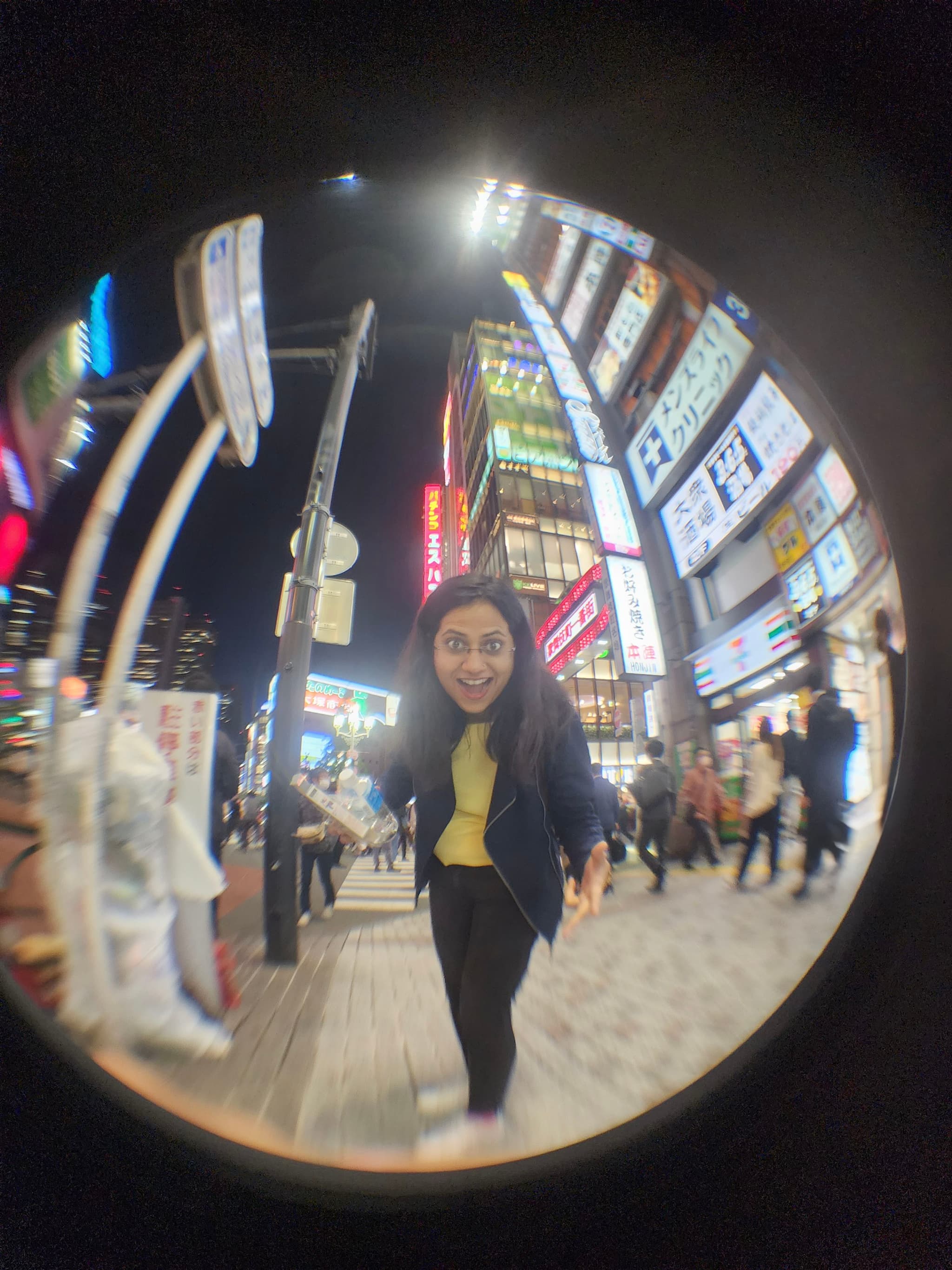 A fisheye lens captures a bustling urban street scene at night, with a woman in the foreground smiling and leaning towards the camera Brightly lit signs and buildings line the street, and several pedestrians are visible in the background