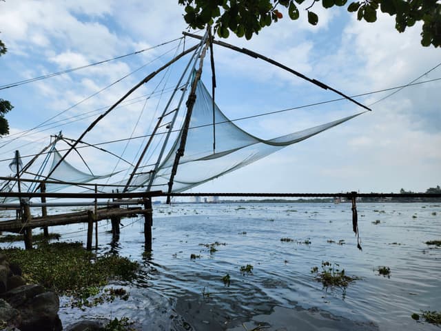 Large fishing nets suspended over a body of water, supported by wooden structures, with a partly cloudy sky and some greenery in the foreground