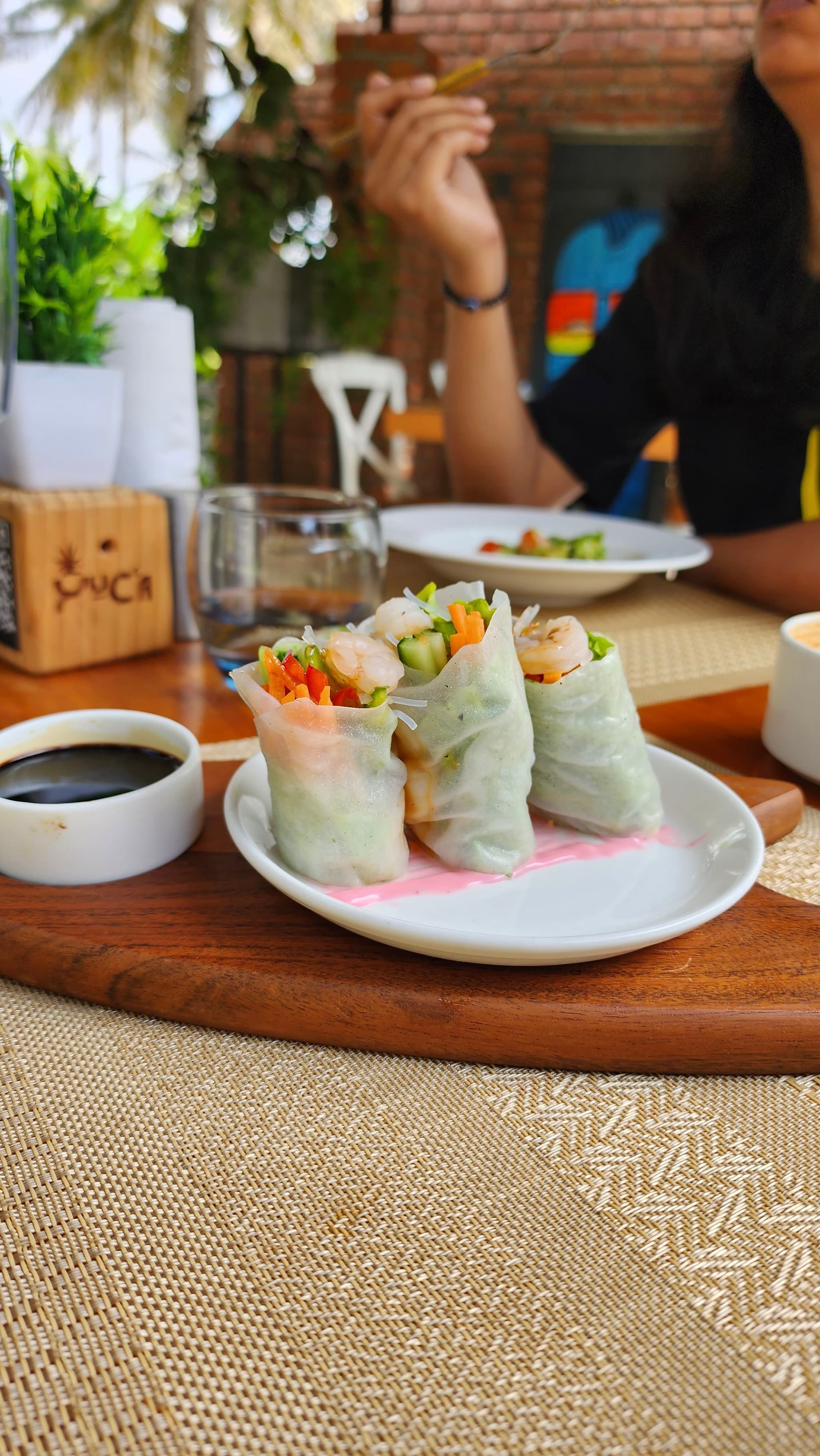 Three fresh spring rolls on a white plate, accompanied by a small bowl of dipping sauce, placed on a wooden board A person is seated at the table in the background, partially visible, with a hand raised holding food The setting appears to be a casual dining environment with a woven placemat and some greenery in the background