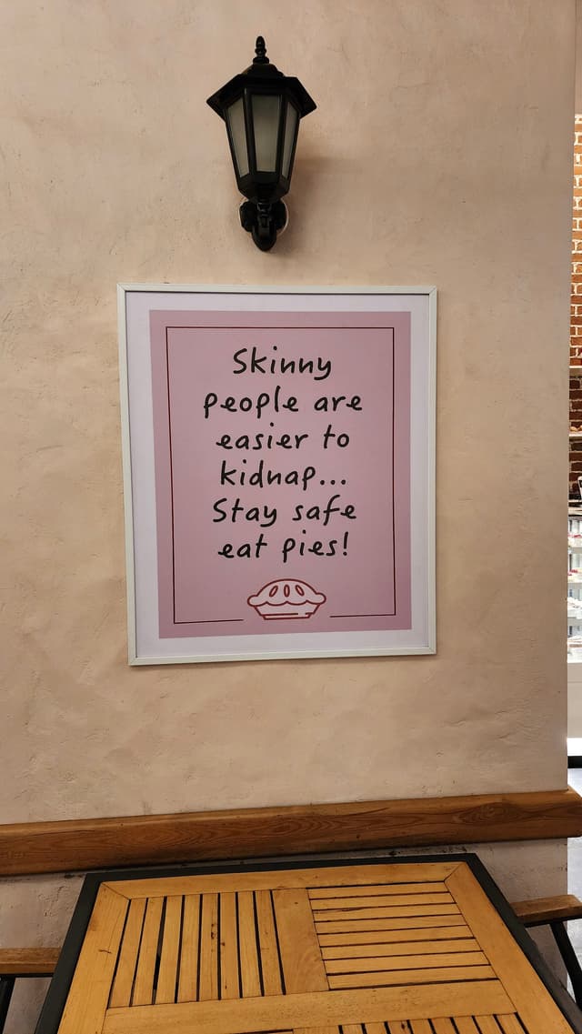 A framed sign on a wall reads, Skinny people are easier to kidnap.. Stay safe eat pies! with a drawing of a pie below the text A black wall-mounted lantern is positioned above the sign, and a wooden table is situated below it