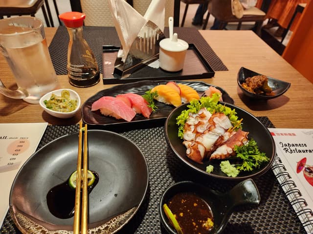 A table set with various Japanese dishes, including sushi, sashimi, a bowl of salad with octopus, a small bowl of pickles, a soy sauce bottle, and a cup of tea Chopsticks rest on a black plate, and a menu is visible on the side