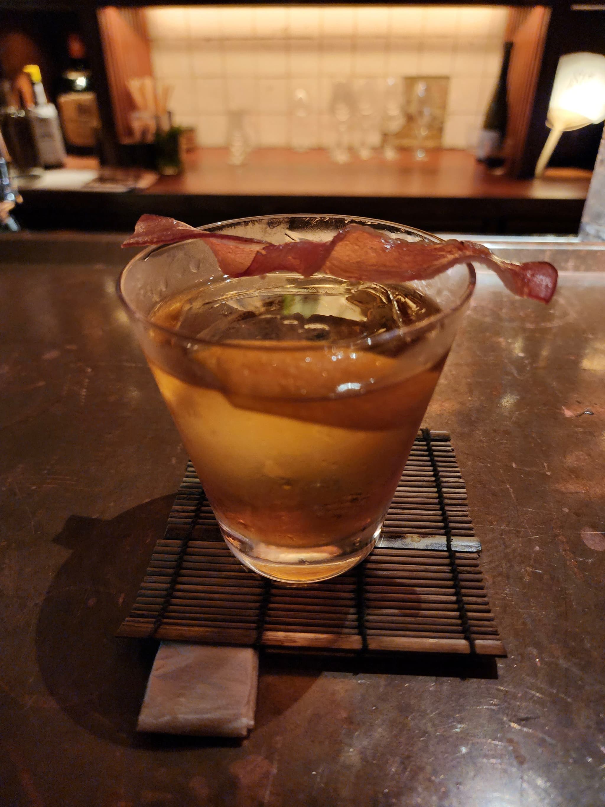 A glass of amber-colored liquid garnished with a twist of citrus peel, placed on a small mat on a wooden surface in a dimly lit setting