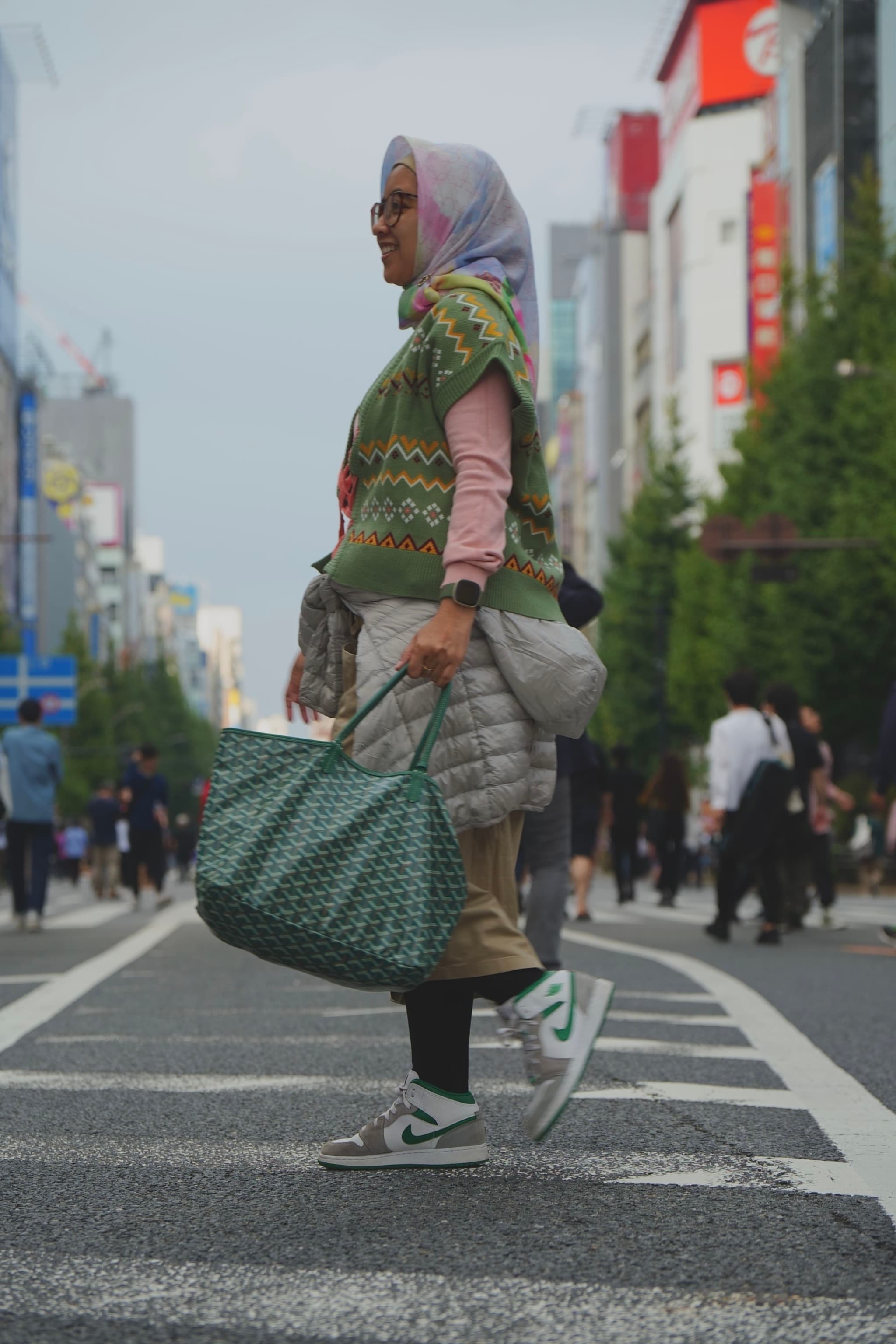 A person wearing a headscarf, glasses, and casual clothing stands in the middle of a city street, holding a large green bag The background features buildings, trees, and other pedestrians