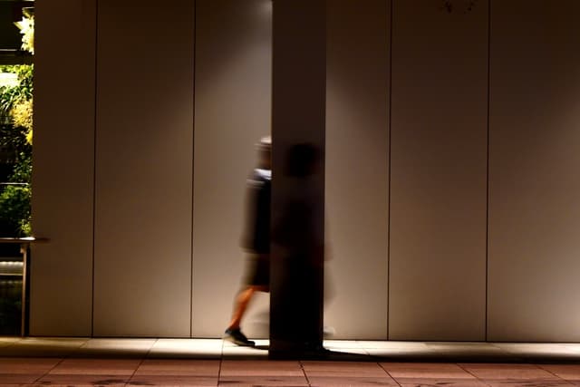 Two blurred figures walking past each other in front of a large, vertical column, with a dimly lit background